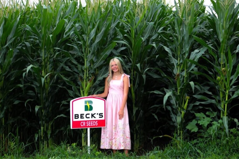 A girl beside a Beck’s CR Seeds sign in a tall cornfield